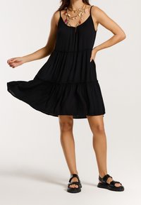 Femme portant une robe noire sans manches à volants, des colliers superposés avec pendentifs et charmes, et des sandales noires, posant avec une main sur la hanche et la jupe tenue vers l'extérieur.