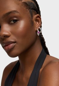 Srebrne kolczyki w kształcie kół mają różowe i niebieskie kamienie szlachetne, prezentując teksturowany design. Modelka ma warkocz i nosi top na halter.