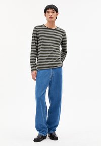 Gestreepte lange mouwen shirt in olijfgroen en grijs, gecombineerd met lichtblauwe wijde jeans en zwarte schoenen, staande op een witte achtergrond.