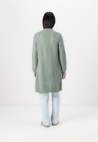 Lång mintgrön cardigan med en texturerad stickad design, sidodelar och en avslappnad passform, tillsammans med ljusblå vida jeans.