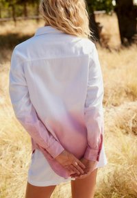 Femme aux cheveux blonds ondulés se tient dans de l'herbe sèche, portant une chemise à manches longues blanche et rose dégradé et un short blanc, les mains jointes derrière le dos.