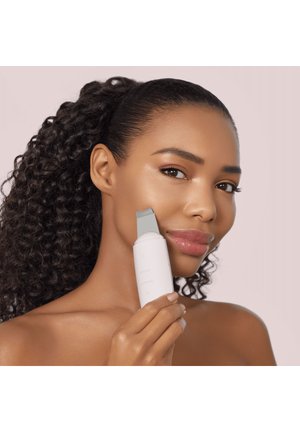 Femme aux cheveux bouclés utilisant un nettoyeur de peau ultrasonique blanc sur sa joue sur un fond clair uni.