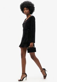 Robe portefeuille en velours noir, à manches longues, de longueur mini, avec un détail à nouer. Associée à des talons aiguilles noirs à lanières et un petit sac à main noir.