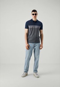 Polo shirt marine avec bloc de couleur gris, manches courtes et col. Porté avec un jean bleu clair et des baskets blanches. Mannequin portant des lunettes de soleil.