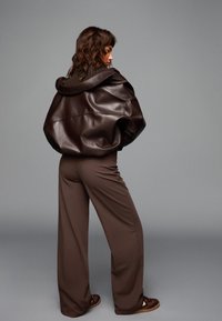 Smeđa kožna oversized jakna s kapuljačom u kombinaciji sa smešnim širokim hlačama. Glatka tekstura, minimalni detalji i opušten kroj.