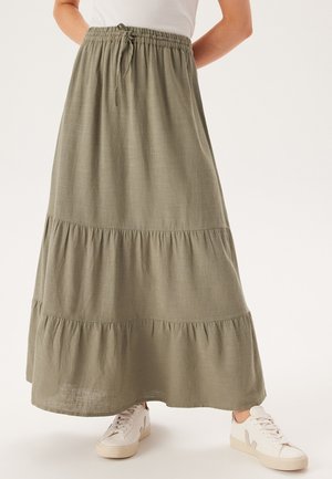 Jupe maxi en vert olive, à volants, avec une taille élastique et un cordon de serrage. Fabriquée en tissu léger et texturé ; associée à des baskets blanches.