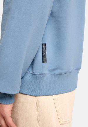 Light blue sweatshirt featuring ribbed cuffs and waistband, with a black label detail on the side. Soft fabric texture, casual design.