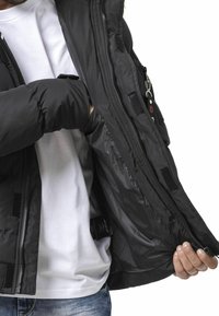 Schwarze Steppjacke mit Reißverschluss vorne. Verfügt über eine glatte Außenseite, mehrere Taschen und ein weiches Futter, das durch die geöffnete Klappe sichtbar ist.