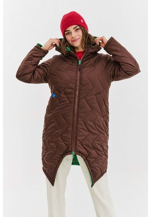 Kobieta poprawiająca kaptur na długim, brązowym pikowanym płaszczu, w czerwonej wełnianej czapce i białych spodniach, stojąca na jasnym tle.