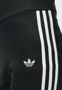 Čierne športové nohavice s tromi bielymi pruhmi po boku a bielym logom Adidas trefoil blízko bedra.
