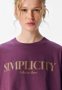 T-shirt en coton violet avec un texte doré en gras portant l'inscription "SIMPLICITY Collectors Team." Encolure ronde et manches courtes. Texture lisse.