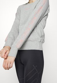Felpa grigia con polsini a costine e scollatura a girocollo, caratterizzata da un logo rosa brillante e dettagli a righe lungo le maniche. Abbinata a leggings neri.