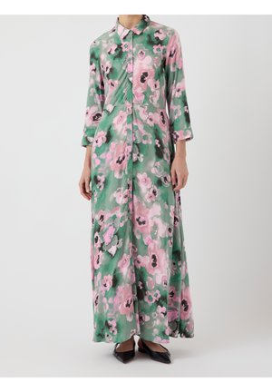 Frau trägt ein langes, geknöpftes Hemdblusenkleid mit rosa-grünem Blumenmuster und dreiviertel Ärmeln, kombiniert mit schwarzen spitzen Ballerinas.