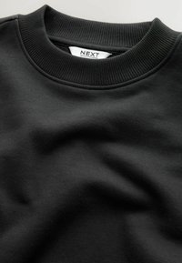 Sudadera negra con cuello redondo acanalado, textura suave y diseño doblado. La etiqueta dice "NEXT", visible en blanco en el collar interior.