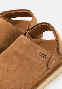 Chaussures UGG en daim marron à enfiler avec une bride arrière ajustable et une semelle beige clair rembourrée, montrées en gros plan.