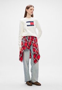 Λευκό φούτερ με ναυτικό και κόκκινο λογότυπο "TOMMY JEANS", επάνω από κόκκινο και μαύρο καρό πουκάμισο δεμένο στη μέση, συνδυασμένο με ανοιχτόχρωμο τζιν παντελόνι.