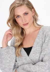 Femeie blondă cu păr ondulat și ochi albaștri, purtând cercei cu cercuri aurii și un pulover moale gri peste un top negru, atingându-și părul.