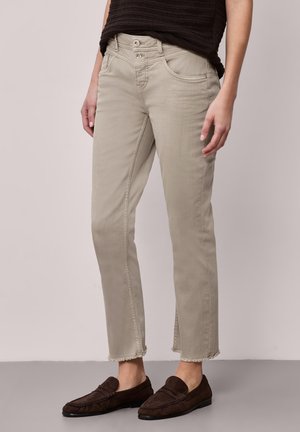 Frau trägt beige, kurz geschnittene Hosen mit rohen Säumen, schwarzes Oberteil und braune Loafers, steht vor einem schlichten Hintergrund.