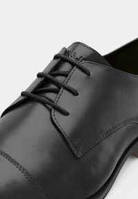 Acercamiento de un zapato de vestir de cuero negro pulido con punta redonda, cordones negros y detalle de costura visible sobre fondo blanco.