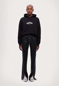 Schwarzes Hoodie mit einem weißen Grafiklogo, kombiniert mit hoch taillierten schwarzen Lederhosen im Schlag und glänzenden silbernen Schuhen. Glatte Textur über die gesamte Fläche.