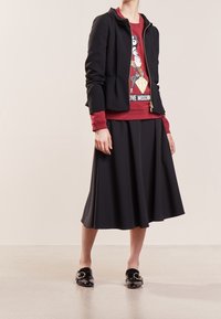 Chaqueta peplum negra sobre un sudadera gráfica roja. Falda negra fluida con pliegues. Zapatos negros de charol con un detalle de hebilla redondeada.