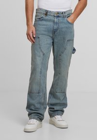 Lockere hellblaue Denim-Jeans mit ausgeprägter Nahtführung und großen Seitentaschen, kombiniert mit weißen Sneakers und einem eng anliegenden weißen Tanktop.
