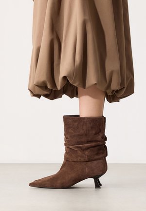 Brown suede mid-calf boots with low angled heels worn with a beige balloon-hem skirt on a light floor and neutral background.
