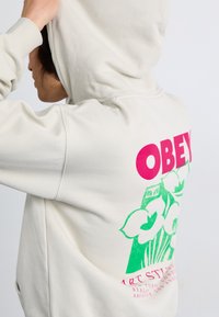 Cream-colored hoodie featuring a large pink "OBEY" logo on the back, with green floral graphics and text detailing at the bottom.