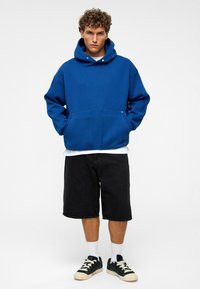 Blauwe hoodie gemaakt van fleece, met een kangoeroezak, metalen drukknoopdetails, en gedragen met zwarte shorts en sneakers.