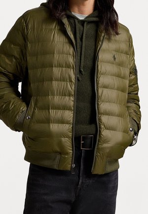 Personne portant une veste matelassée vert olive non fermée sur un pull à capuche vert foncé et un pantalon noir avec une ceinture noire.