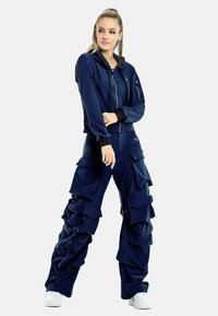 Mörkblå hooded tracksuit med dragkedja, som har lösa cargobyxor med flera fickor och volangdetaljer, i kombination med vita sneakers.