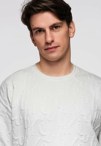 Pull-over blanc avec motif texturé en lettres surélevées. Présente un col rond et des poignets côtelés avec une texture douce tricotée.