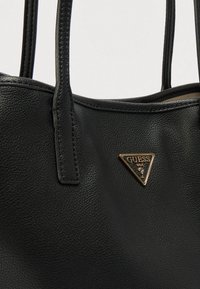 Melna sintētiskā tote soma ar fakturētu virsmu, divām rokturiem un zelta trijstūra formā izcelti logotipas akcenti. Vienkāršs un praktisks dizains.