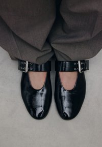 Zwarte lakleren schoenen met een ronde neus en uitsparingen aan de zijkanten. Verstelbare enkelbandjes met metalen gespen. Gedragen met grijze stoffen broeken.