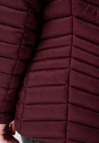 Doudoune bourgogne présentant une texture matelassée, des coutures horizontales et inclinées, ainsi qu'un tissu doux et brillant avec une finition lisse.
