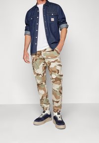 Denim overhemd met knoopsluiting, witte T-shirt en beige-groene camouflage cargo broek. Marineblauwe sneakers met beige accenten. Casual fit.