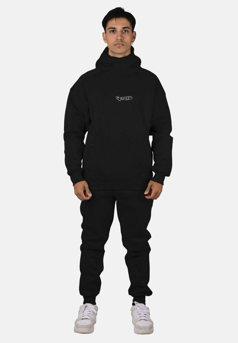 Sudadera con capucha y pantalones joggers negros de mezcla de algodón, con cuello alto y logo bordado en el pecho. Zapatillas blancas completan el conjunto.