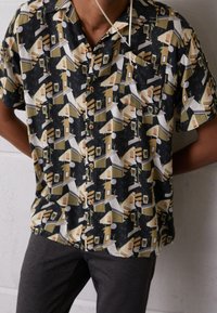 Camicia a maniche corte con bottoni, caratterizzata da un motivo geometrico in nero, crema e oliva. Il tessuto sembra leggero e traspirante.