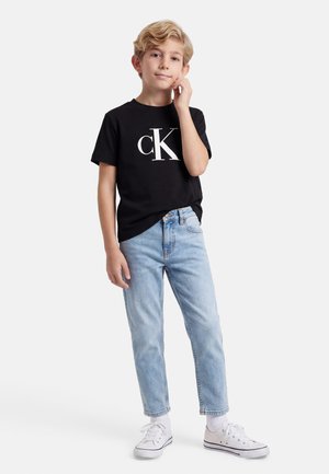 Blonder Junge steht mit der Hand nahe am Gesicht, trägt ein schwarzes T-Shirt mit CK-Logo, helle blaue Jeans, weiße Socken und weiße Turnschuhe vor weißem Hintergrund.