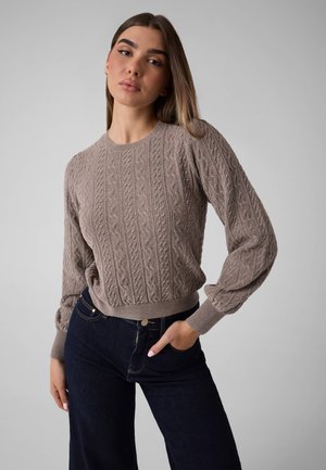 Femme aux longs cheveux bruns portant un pull torsadé taupe et un jean bleu foncé, debout avec une main dans la poche devant un fond uni.