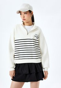 Wit sweatshirt met ronde hals, zwarte strepen, ritskraag, geborduurd "Los Angeles Club" logo, gecombineerd met een zwarte gelaagde rok en een pet.