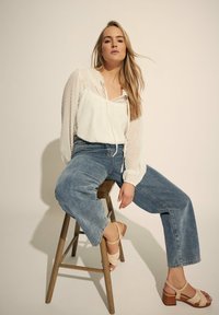 Blusa bianca strutturata con colletto regolabile, abbinata a jeans a gamba larga blu chiaro e sandali con plateau beige, seduta su uno sgabello di legno.