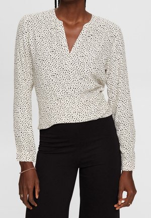 Vrouw draagt een witte blouse met lange mouwen met kleine zwarte stippen en een zwarte broek, toont bovenlichaam en armen.