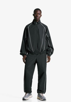 Ensemble de sport noir avec des finitions blanches, un col haut, des poignets élastiques et un pantalon assorti. Le modèle porte des baskets grises.