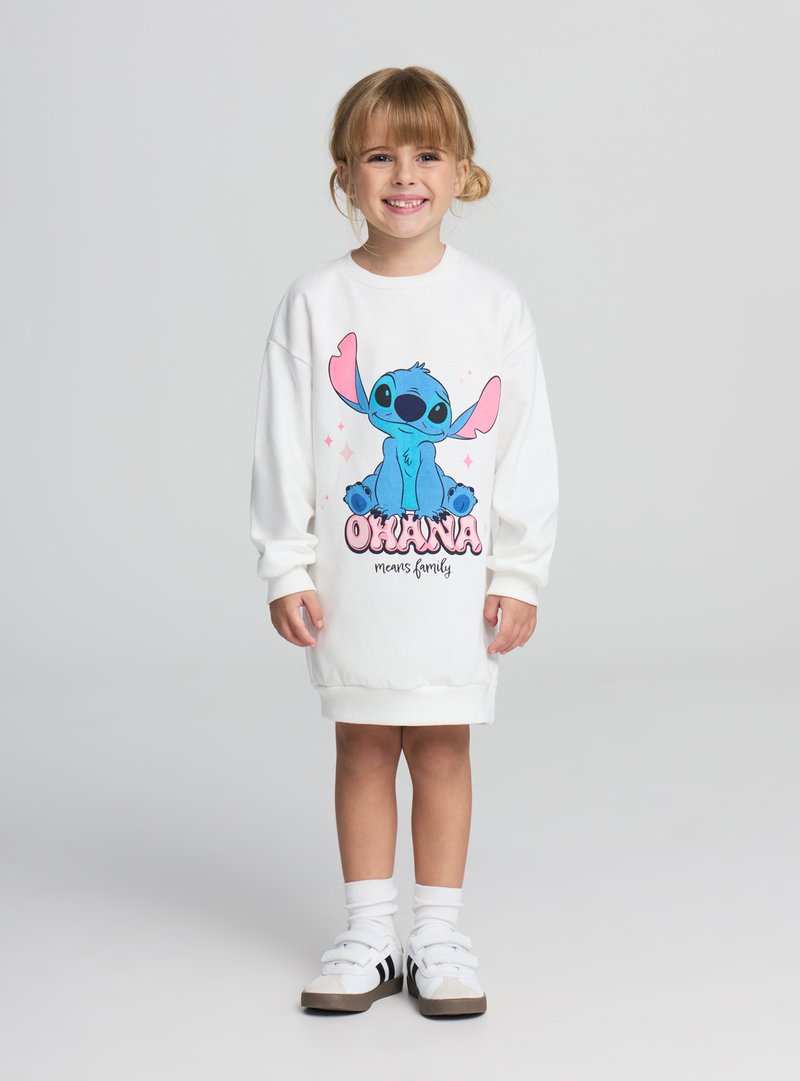 Abito oversize bianco con un personaggio dei cartoni animati blu con grandi orecchie, accompagnato dal testo "Ohana means family" in rosa e nero.