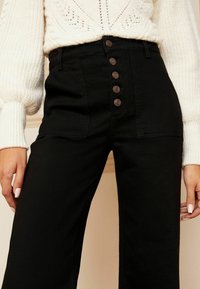 Pantalones negros de talle alto con cierre frontal de botones, tela texturizada y bolsillos laterales, combinados con un suéter de punto color crema.