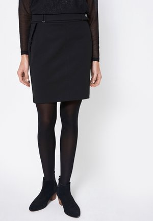Svart blyertskjol med texturerad yta, med en sidobesparing, bältad midja och enkel knappstängning, i kombination med svarta strumpbyxor och ankelskors.