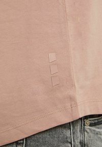 T-shirt rose clair avec une texture douce, manches courtes et trois accents carrés dans une teinte légèrement plus foncée près de l'ourlet.