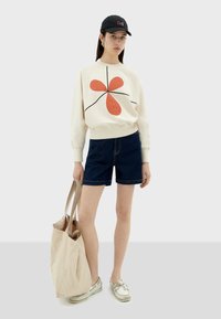 Sweatshirt crème avec un design graphique rouge, shorts en denim foncé et sac fourre-tout beige. Des chaussures blanches complètent ce look décontracté.