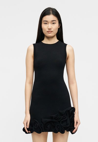 Γυναίκα με μακριά μαύρα μαλλιά που φοράει μαύρο αμάνικο φόρεμα με βολάν στο τελείωμα, στέκεται μπροστά σε απλό ανοιχτόχρωμο φόντο.